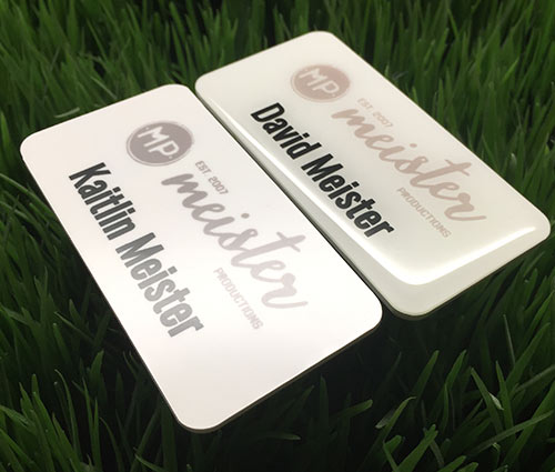 Photo of two plastic nametags. One with epoxy coating. Design for Meister Productions.