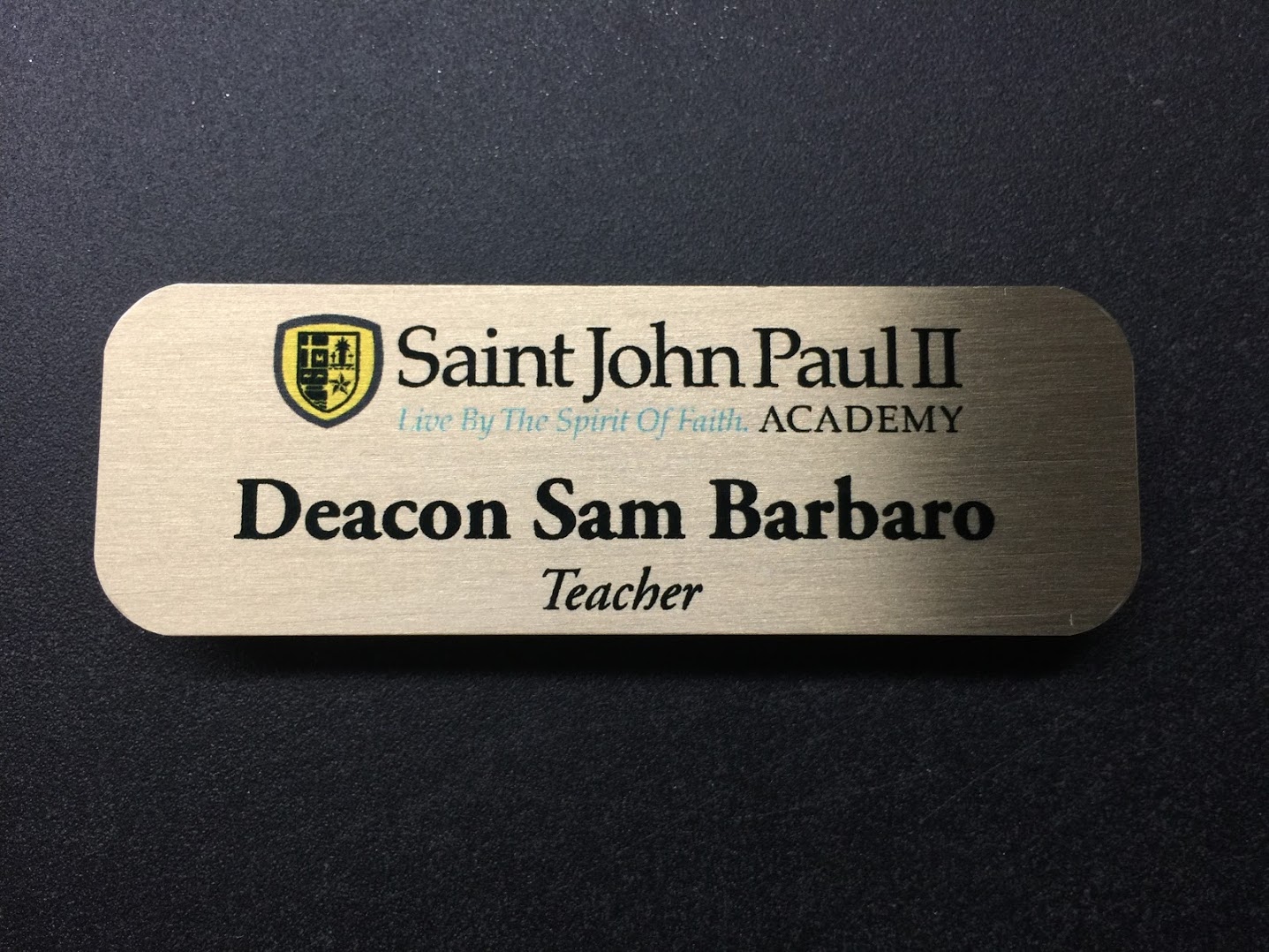 A brushed gold nametag. The design is for Saint John Paul II Academy.